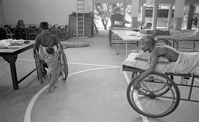Ein körperlich behinderter Mann im Rollstuhl wird von seinem Sohn getragen; ein anderer Mann mit dem gleichen Schicksal beobachtet dies im Zentrum für die Rehabilitation der Gelähmten, CRP in Savar, Dhaka, Bangladesch. 2005.